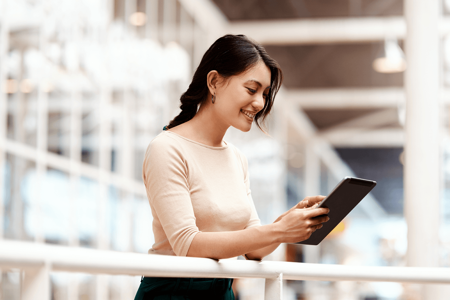 A woman with long dark hair tied back is smiling while using a tablet, standing in a modern indoor setting with glass walls. She is dressed casually in a beige top, appearing engaged and focused, suggesting a professional context such as hiring an agency or exploring digital PR services.