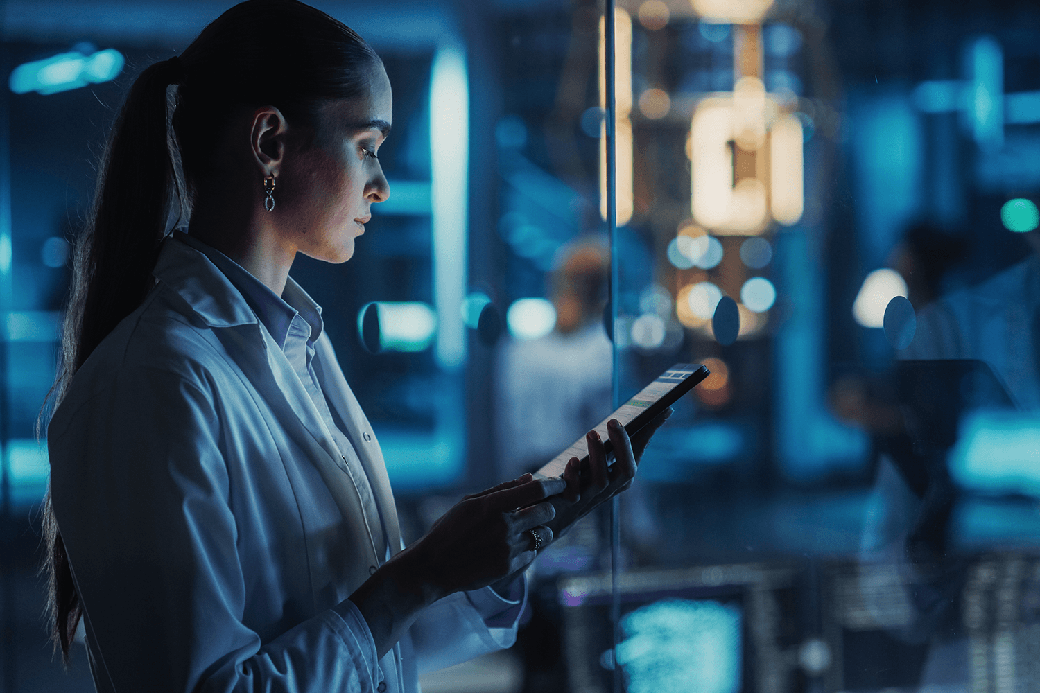 Woman in a white lab coat using a tablet in a dimly lit high-tech laboratory, with blue ambient lighting and reflections on glass panels creating a futuristic medical research setting.