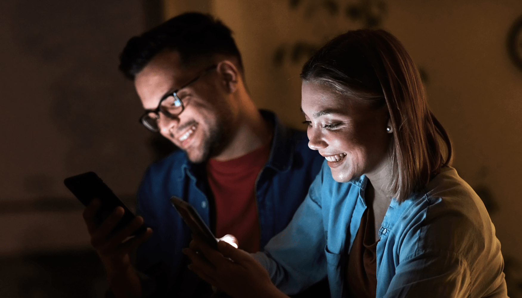 Two people smiling at their phones in a dim setting illustrate how emotionally engaging content captures attention and encourages real-time sharing behavior on social media.
