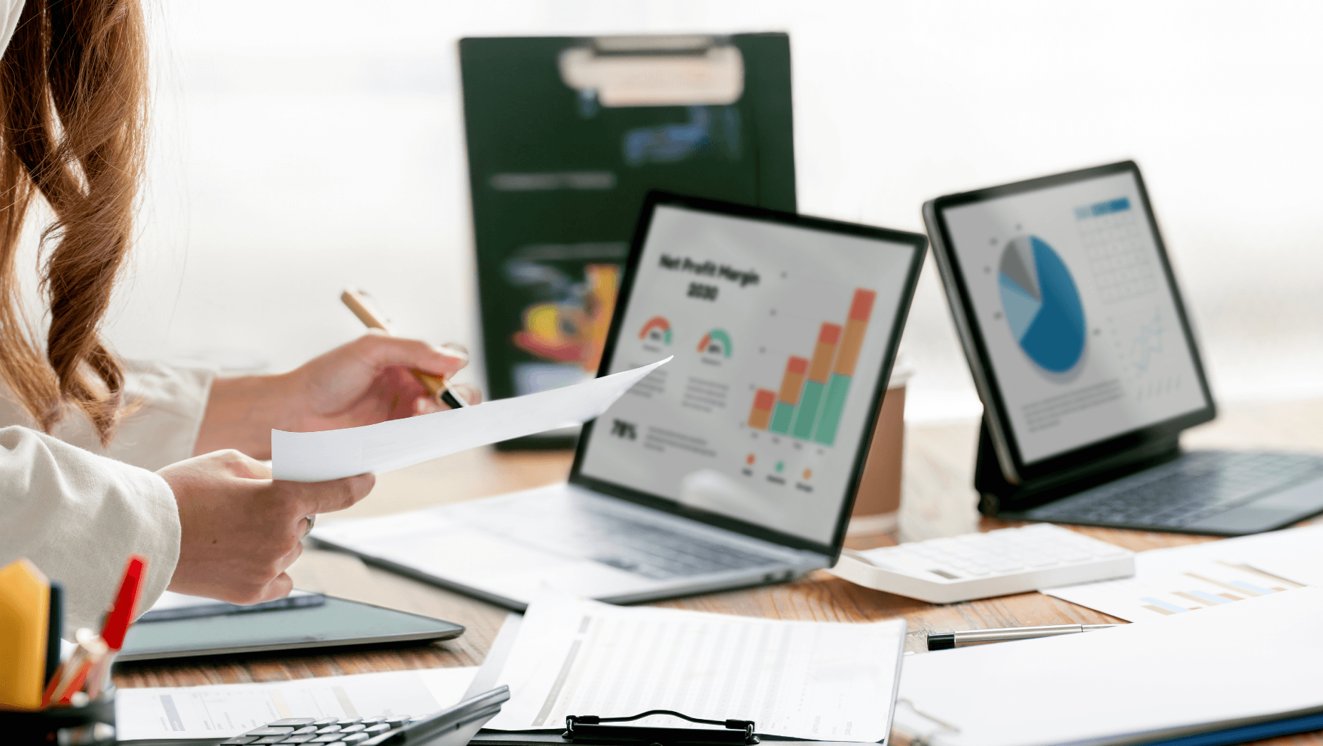 Woman at a desk reviewing profit-margin bar charts and a pie chart on a laptop and tablet, analyzing business performance data