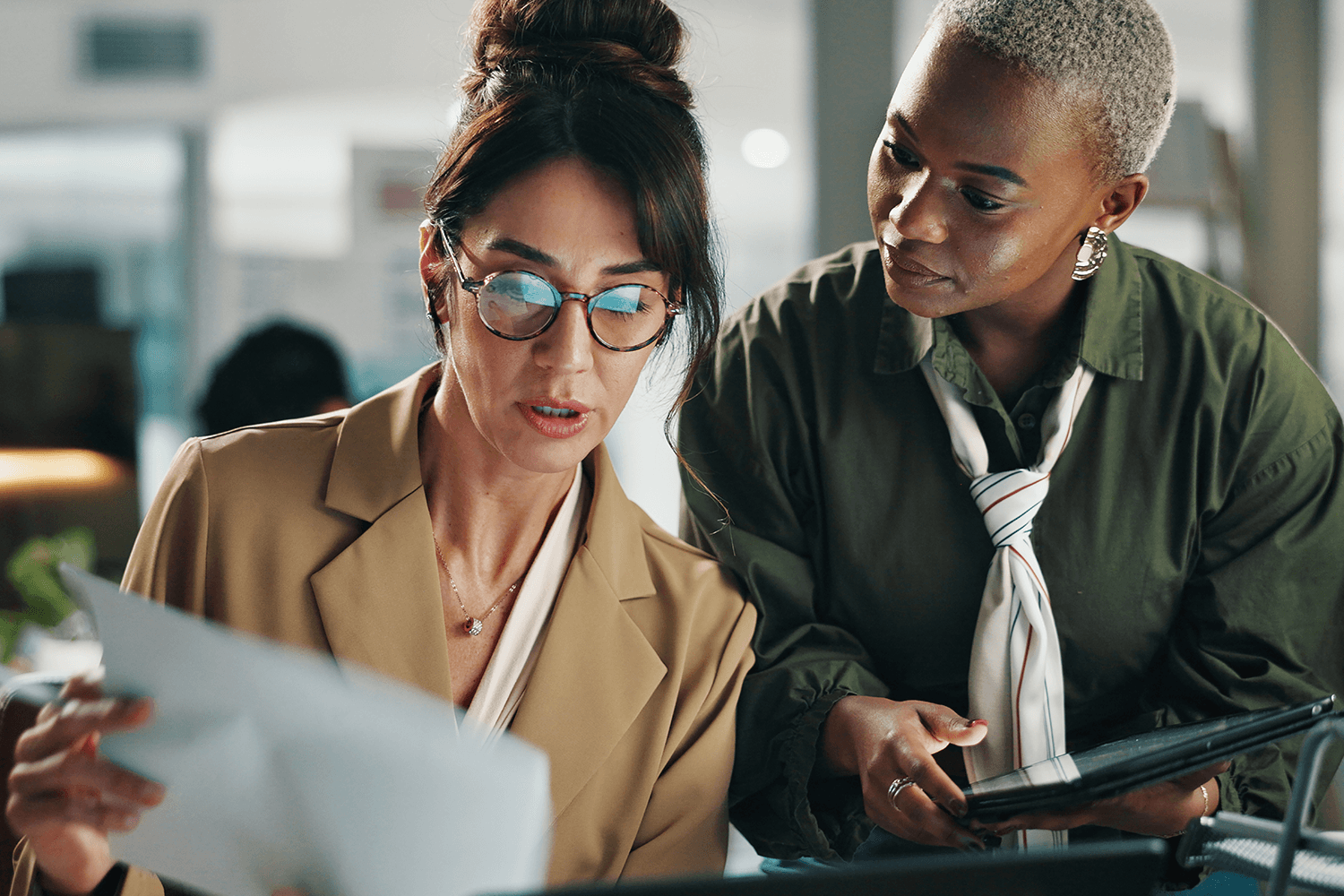 Two businesswomen reviewing a document and tablet together in an office, collaborating on a digital PR or marketing strategy