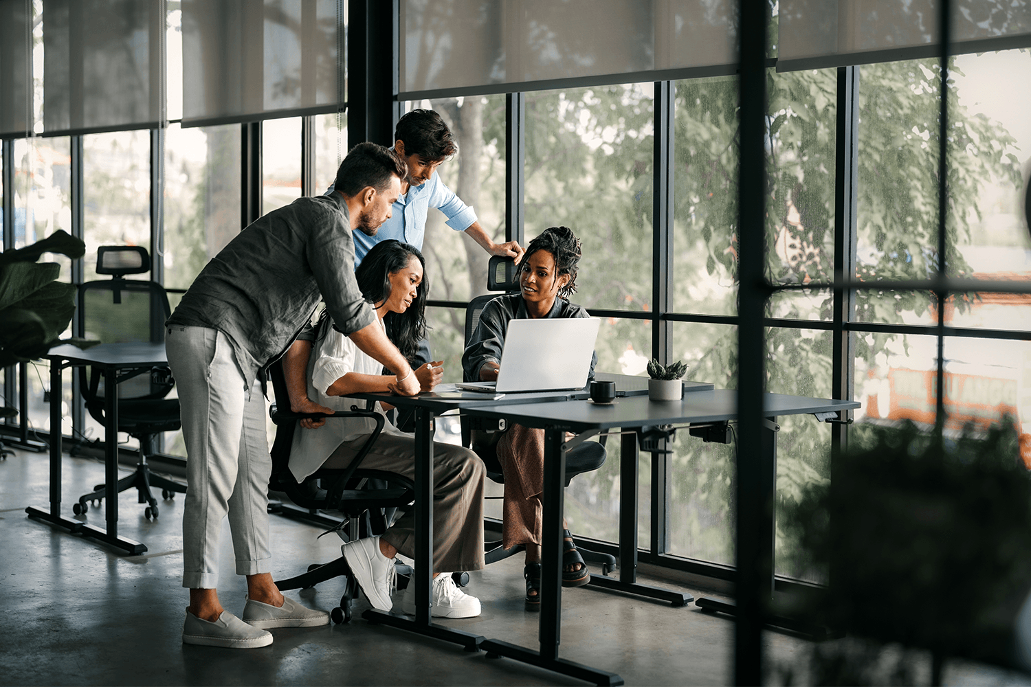 Photograph of four colleagues gathered around a laptop at a desk in a bright, modern office with floor-to-ceiling windows and greenery outside. Two people are seated and two stand behind them, all looking at the screen and collaborating.