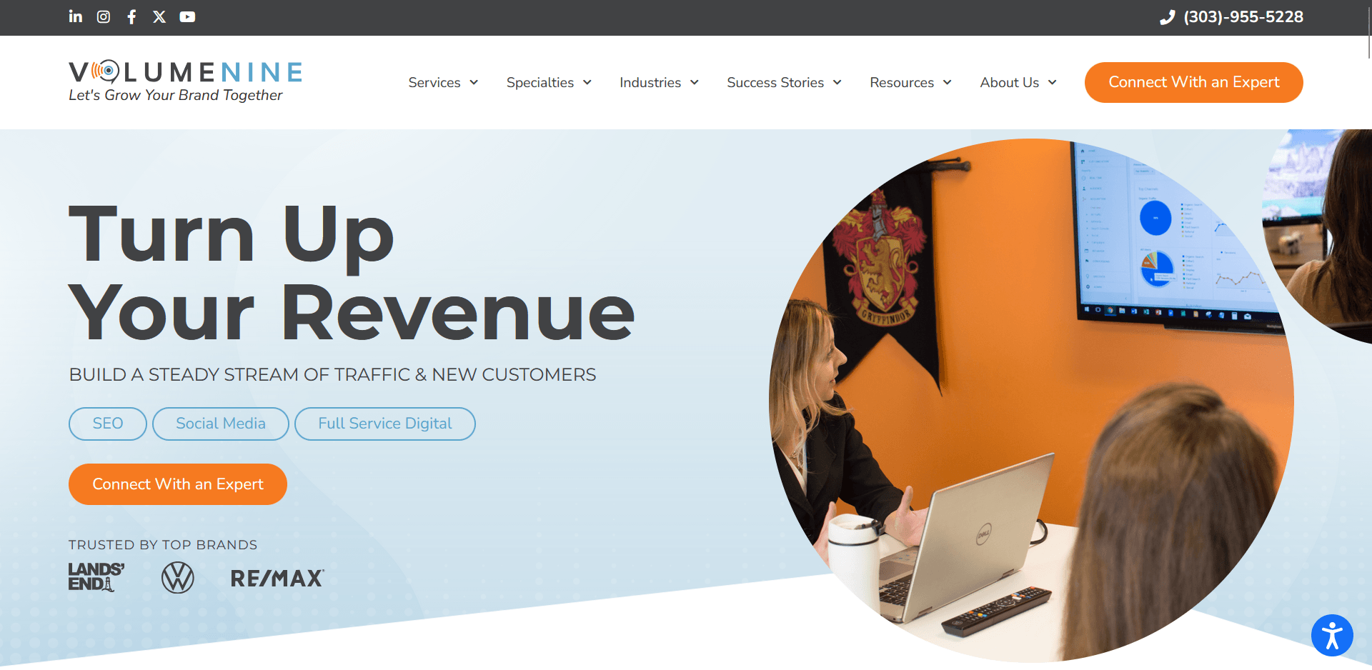 A marketing agency homepage features the bold headline “Turn Up Your Revenue” with subtext promoting increased traffic and customer growth. To the right, a circular photo shows two people in a meeting room viewing analytics on a large screen, with an orange “Connect With an Expert” button below the text.
