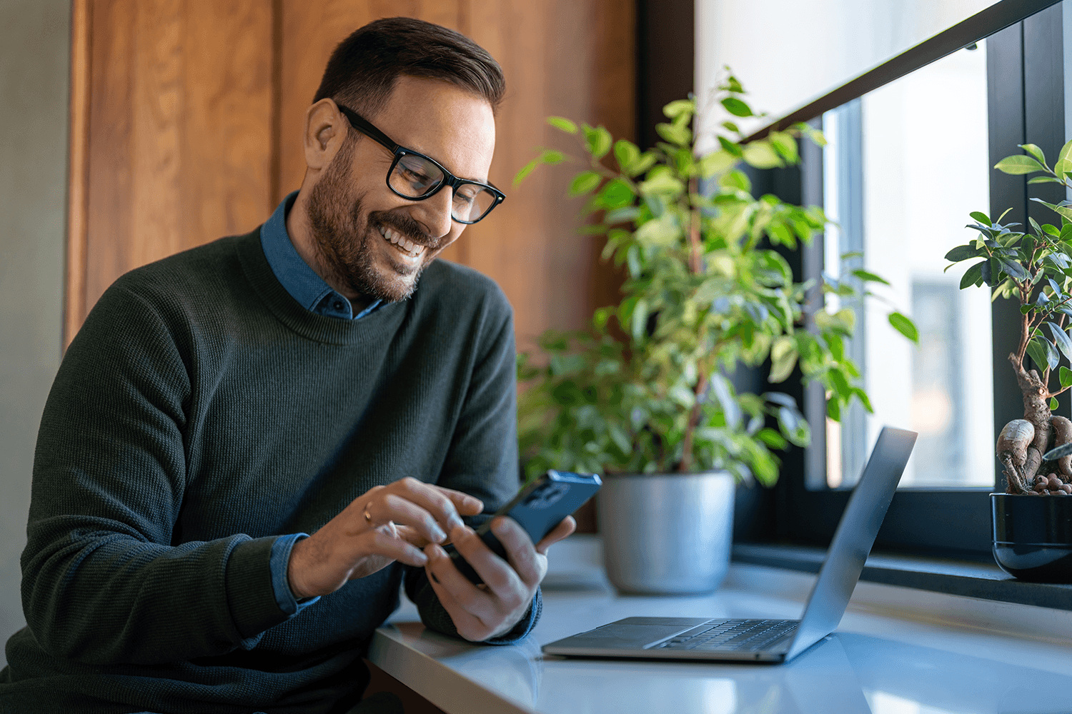 Marketer smiling as he checks his phone next to a laptop, tracking how often ChatGPT and other AI assistants cite his brand.
