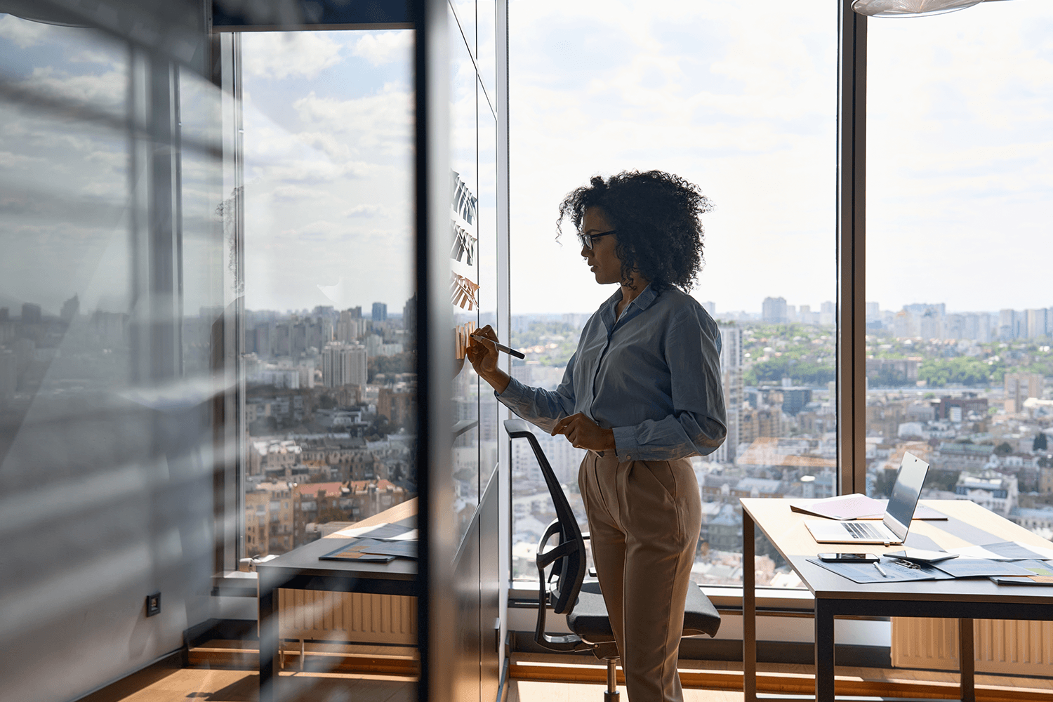 Woman writing on a glass wall in a high-rise office overlooking a city skyline, with a laptop and notes on a desk nearby.